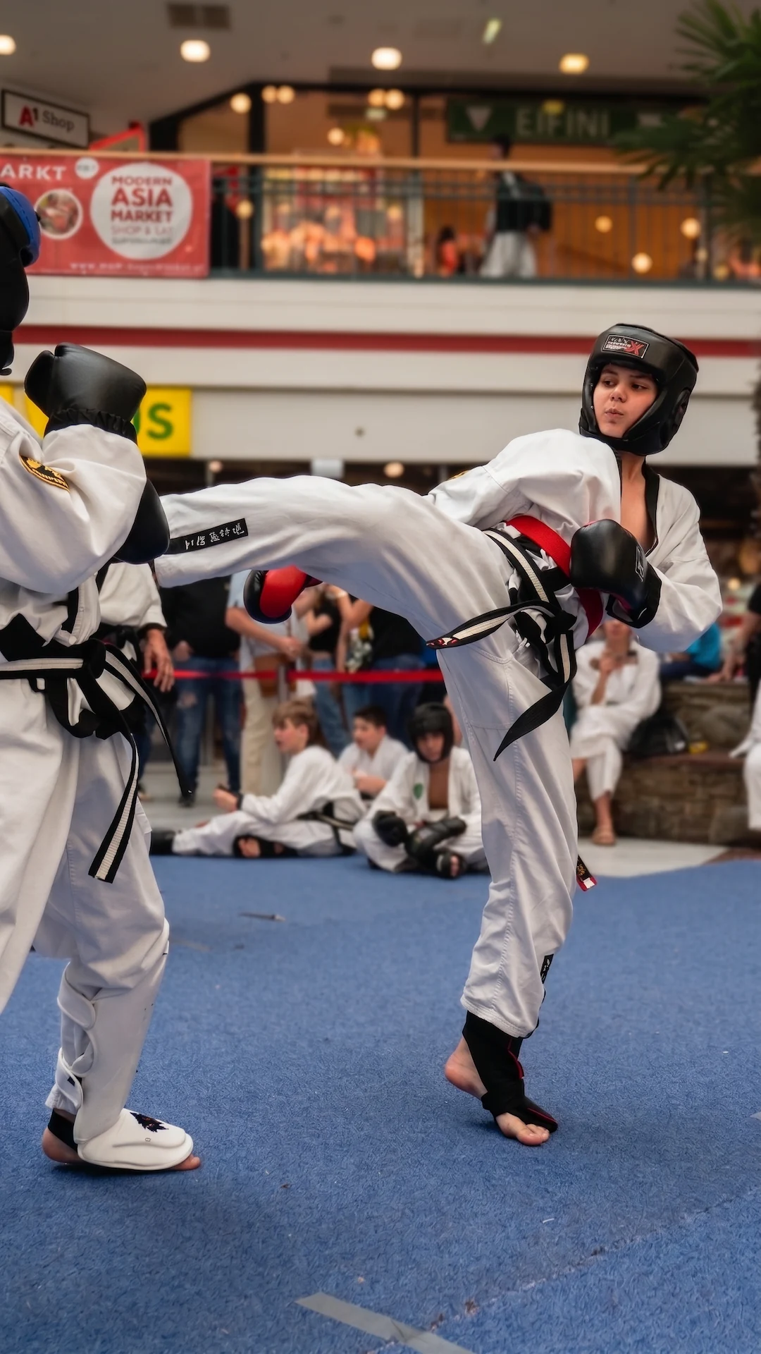 Taekwondo Liga-Turnier für Erwachsene im Einkaufszentrum: Ein Kämpfer zeigt einen hohen Kick auf blauer Matte.