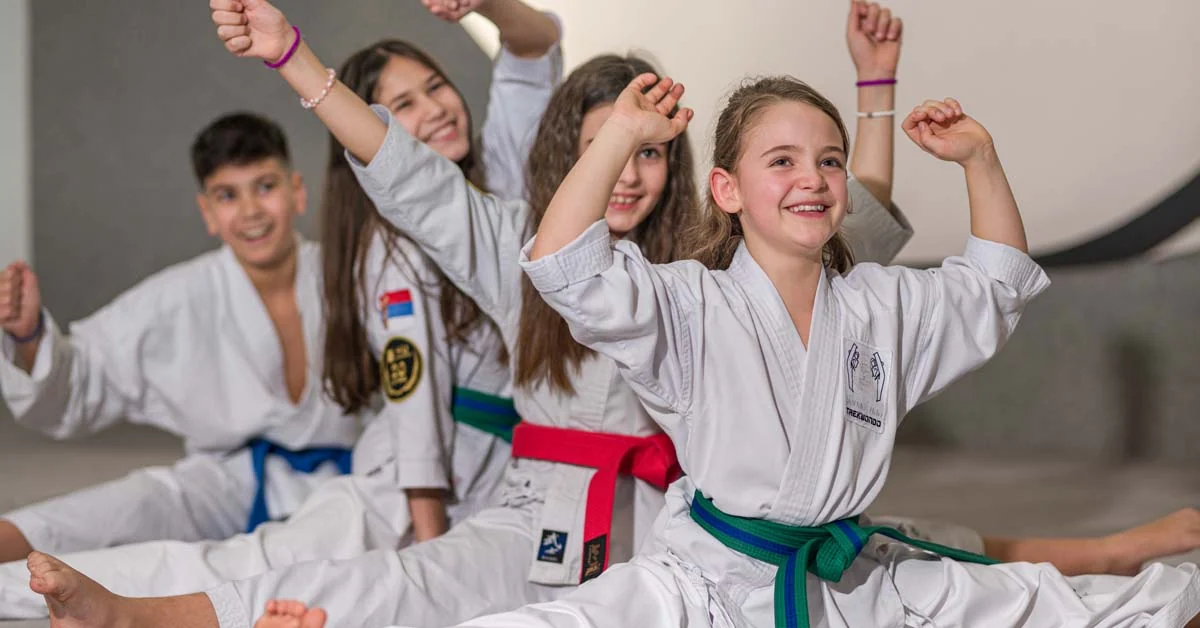 Vier lachende Kinder im Karate-Gi beim Dehnen im Spagat während des Kampfsport-Trainings im Dojo.