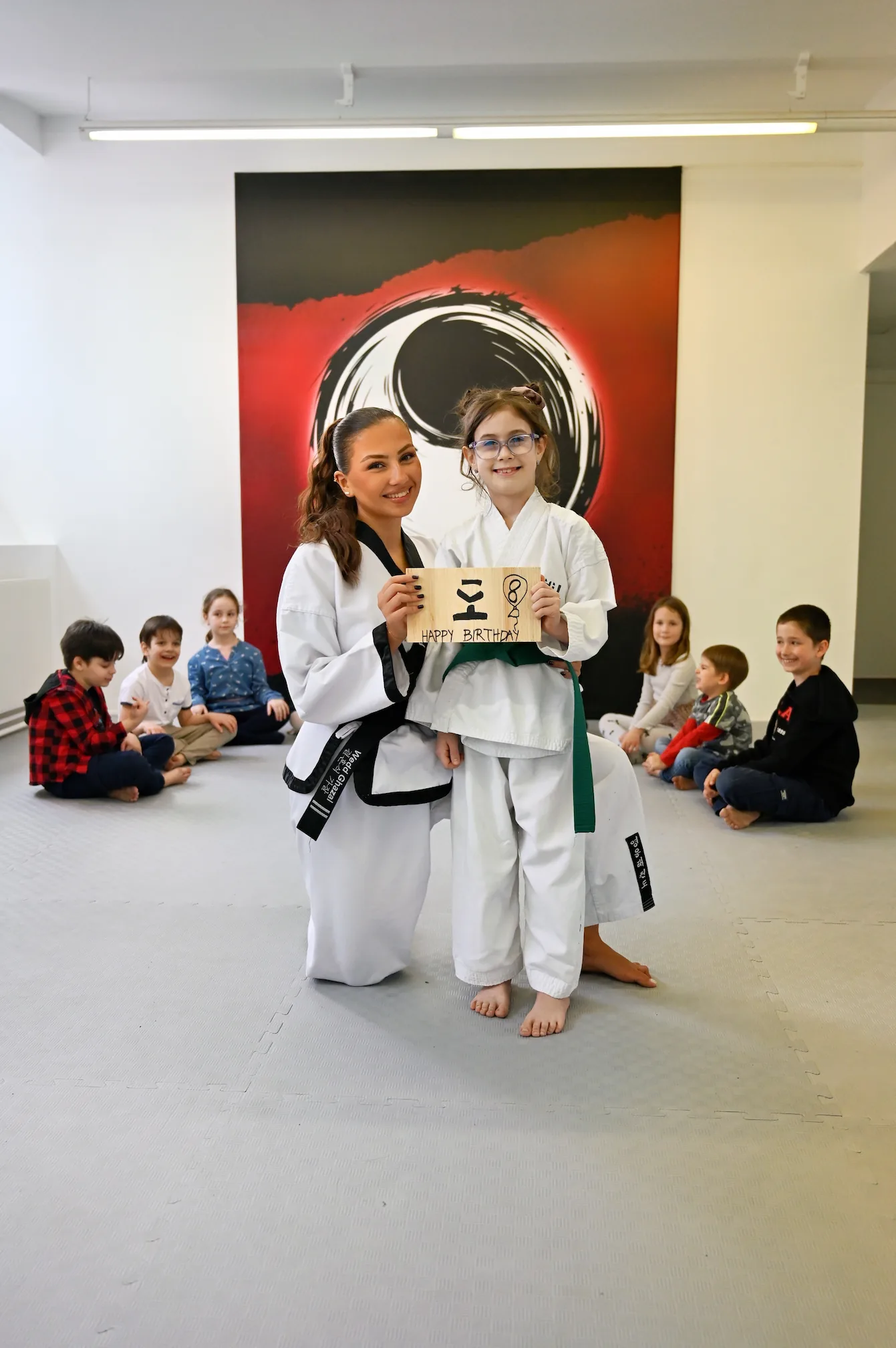 Kindergeburtstag bei YU Taekwondo: Eine Trainerin mit Schwarzgurt und ein Mädchen im Dobok mit Grüngürtel halten ein Geburtstags-Bruchtestbrett.