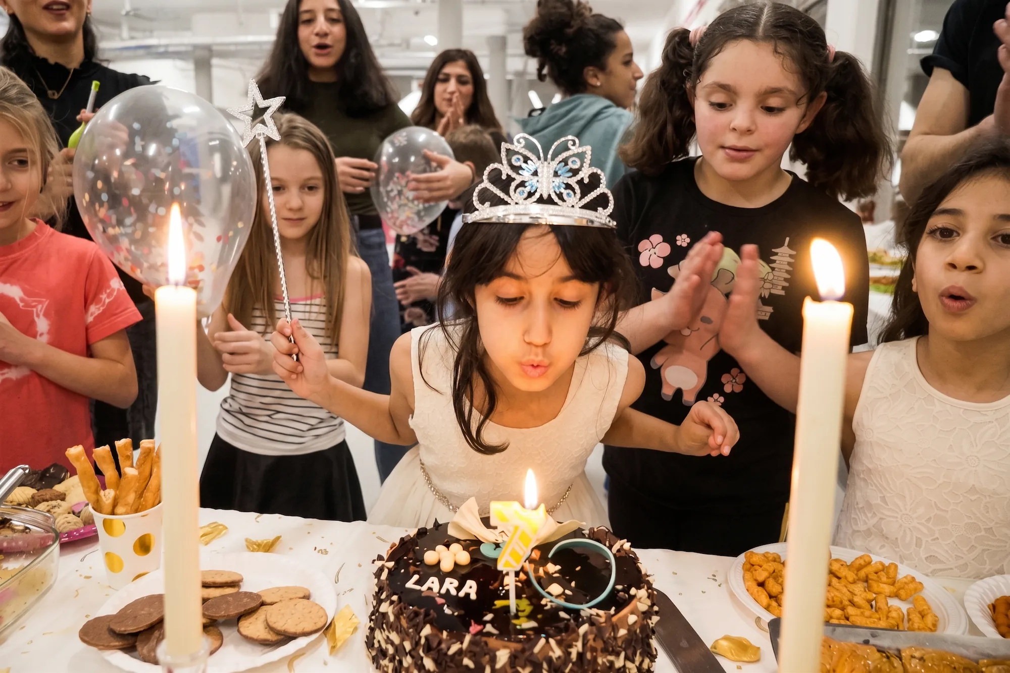 Kinder feiern einen Taekwondo-Kindergeburtstag: Ein Mädchen mit Krone pustet die Kerze auf ihrer Schokotorte mit der Aufschrift „Lara“ aus, während Freunde in einem hellen Trainingsraum zuschauen und klatschen.