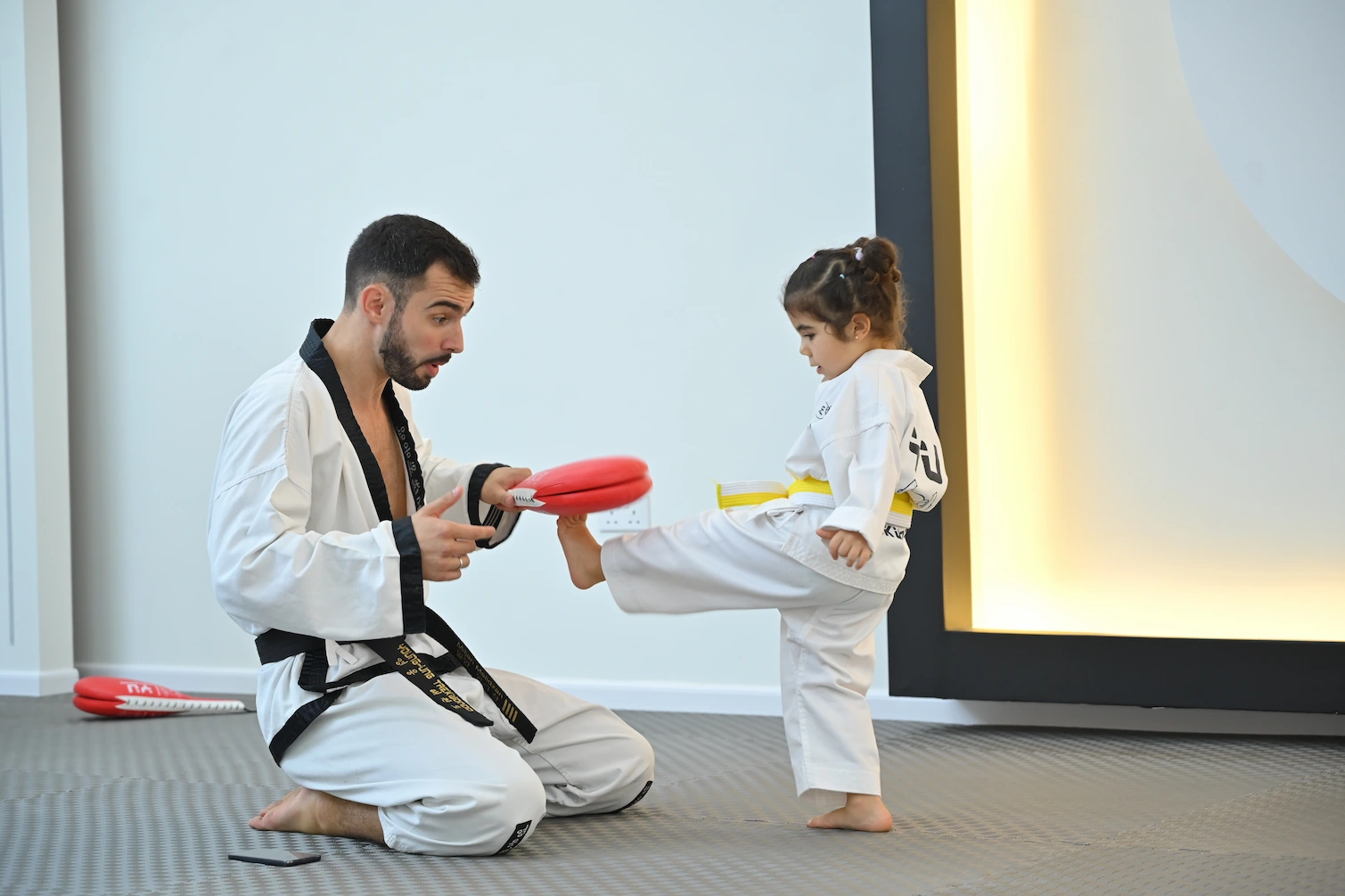 Kleines Mädchen beim Taekwondo-Kindertraining führt Tritt gegen Pratze aus, die ein Trainer hält.