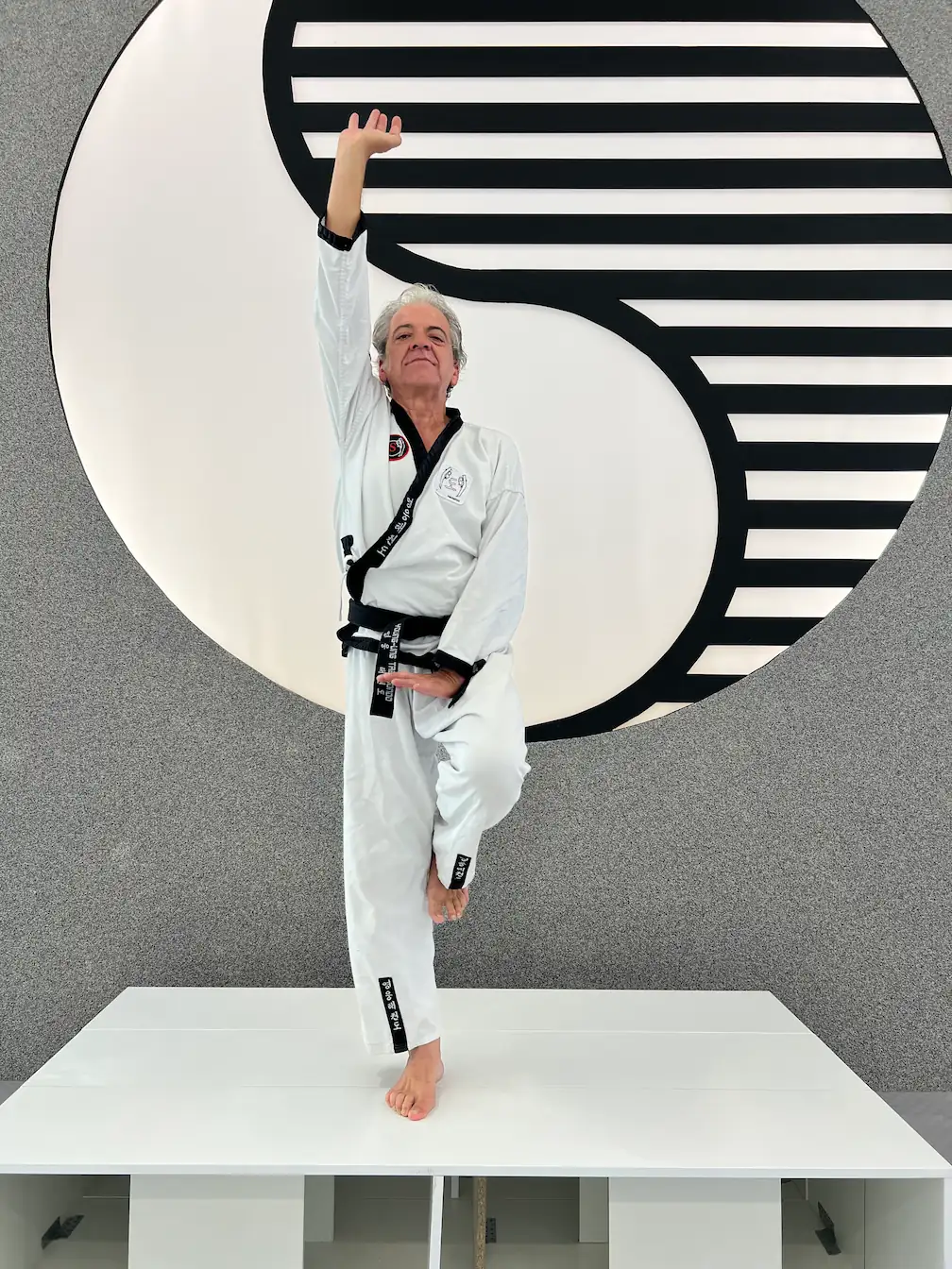 Taekwondo-Senior mit Schwarzgurt übt eine balancierte Poomsae-Technik im Training vor einem Yin-Yang-Logo.