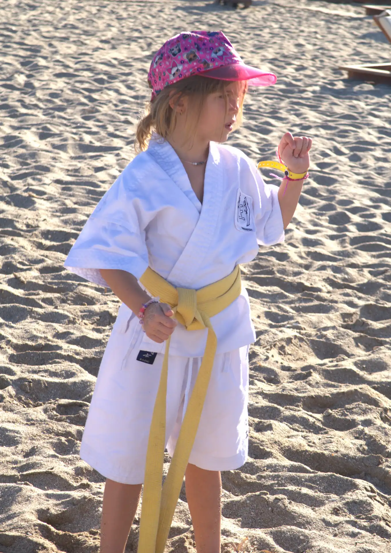 Kind im Judo-Anzug mit gelbem Gürtel und bunter Kappe übt Karate-Posen am Strand eines Sommercamps.
